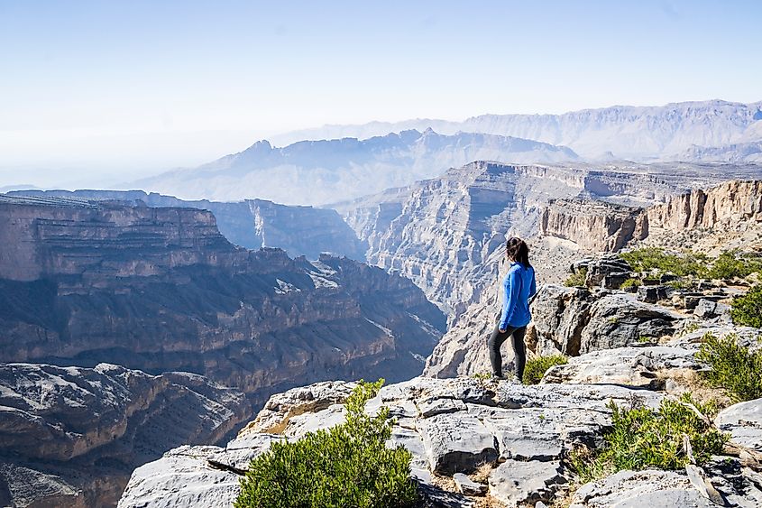 A solo woman hiking through the rugged terrain of Jebel Shams, overlooking the dramatic cliffs of Wadi Ghul, also known as the Grand Canyon of Arabia, in Oman.