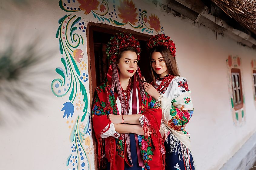 Two Ukrainian women in traditional clothing with floral wreaths in front of a decorated hut. Image Credit BONDART PHOTOGRAPHY via Shutterstock.